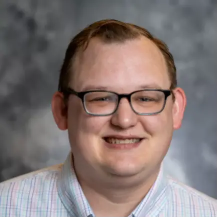 Smiling person wearing black-framed glasses and a light striped button-down shirt against a gray studio backdrop.