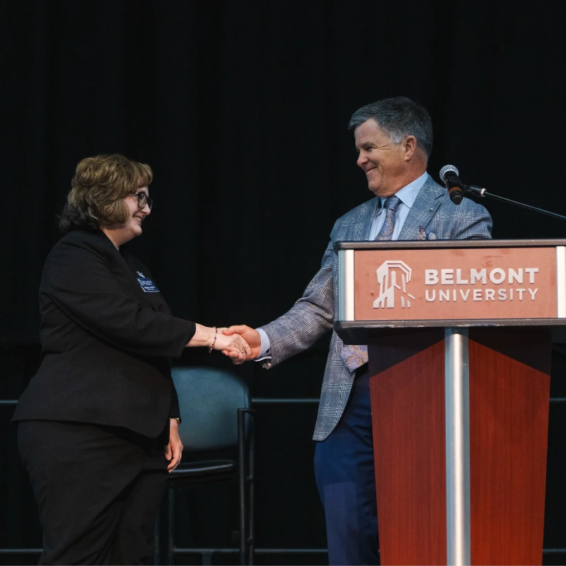 Woman and man shaking hands near a Belmont University podium, both smiling.