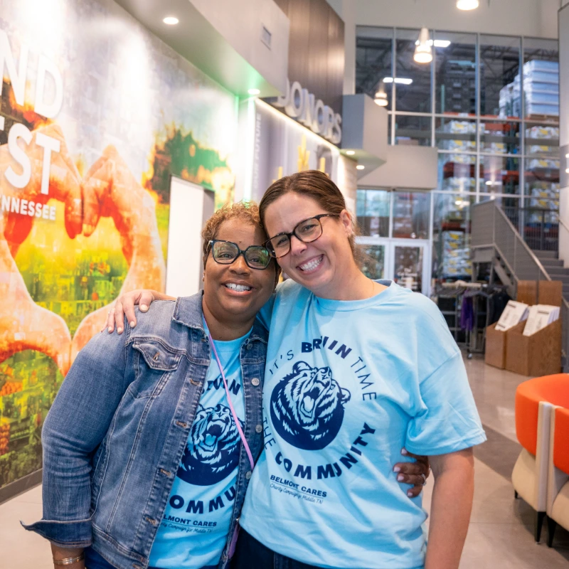 Two happy women wearing "Belmont Cares" community t-shirts, embracing and smiling at a volunteer event.