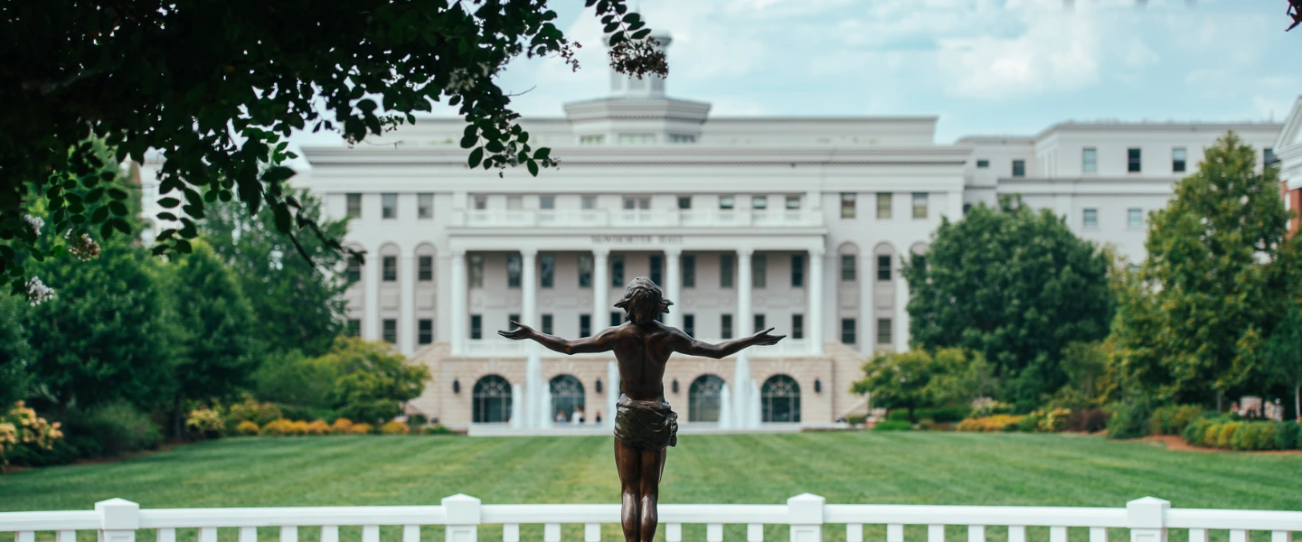 Bronze statue of Christ with open arms on a green lawn, facing a grand white building with classical columns.