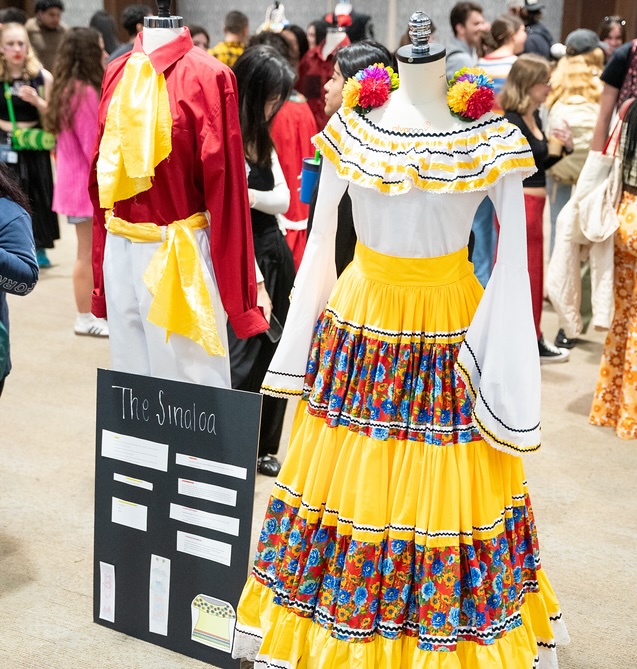 Colorful traditional Latin American costumes on display at a cultural event, featuring a vibrant yellow tiered dress with floral patterns and white ruffled top alongside red and yellow garments, with an informational placard labeled "The Sinaloa" in the foreground and attendees visible in the background.