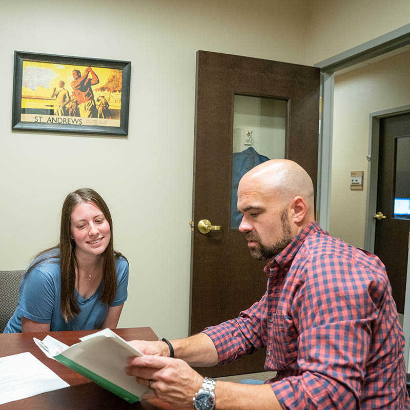 A professor reading from a book in his office while she sits across from him during his office hours