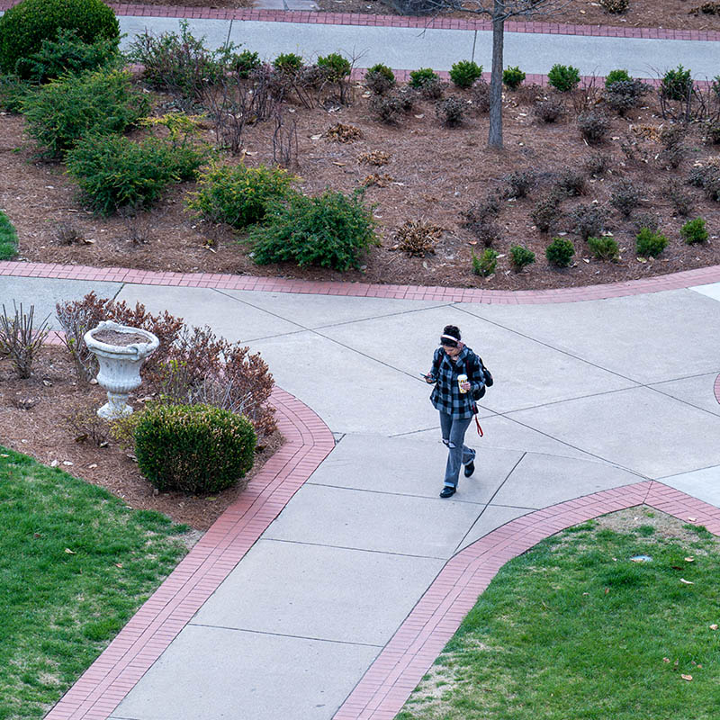 Student wearing headphones while walking on campus while looking at her phone and holding a cup of coffee