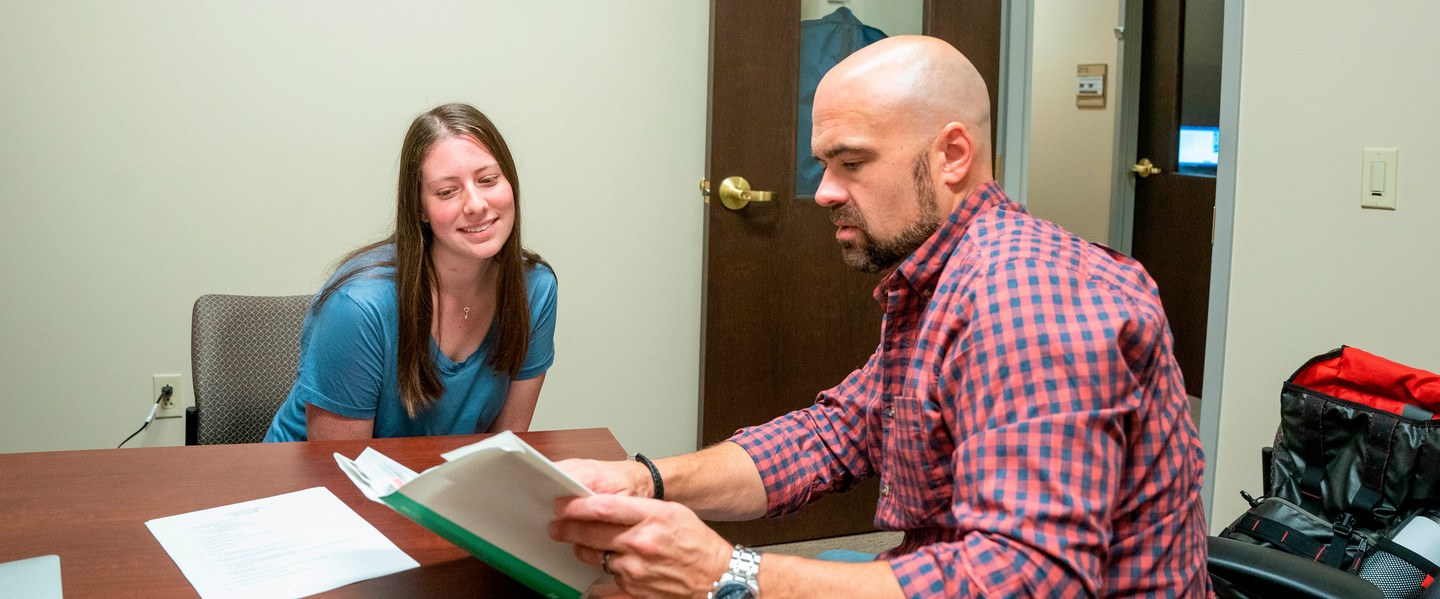 A professor reading from a book in his office while she sits across from him during his office hours