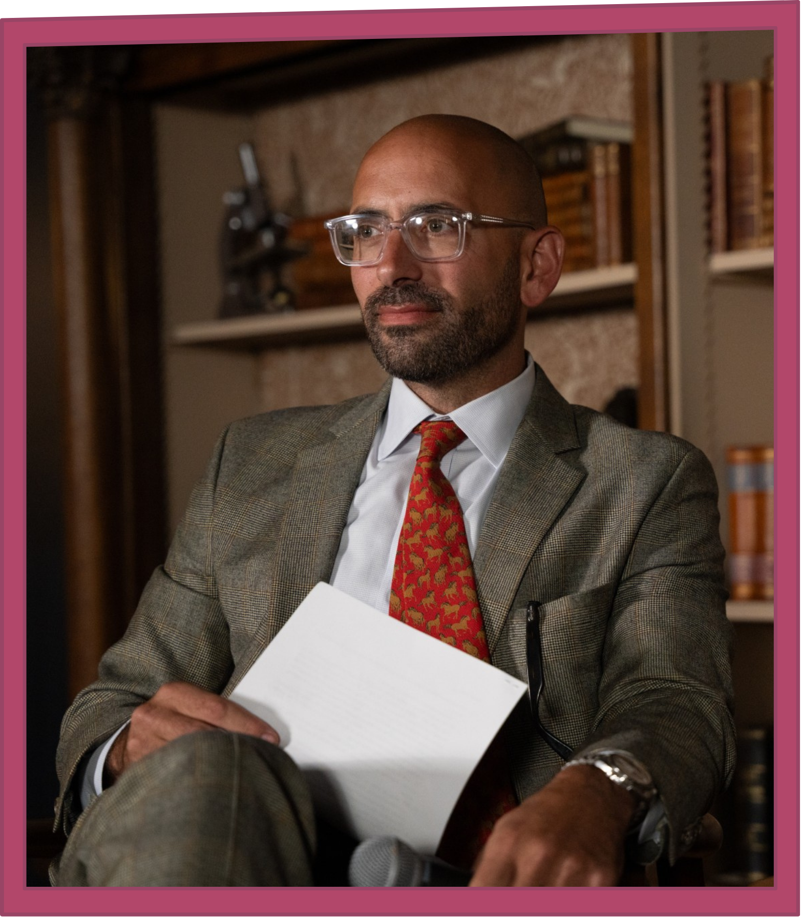 Bald man wearing clear-framed glasses and a suit with a red patterned tie, seated and holding a sheet of paper in front of bookshelves.