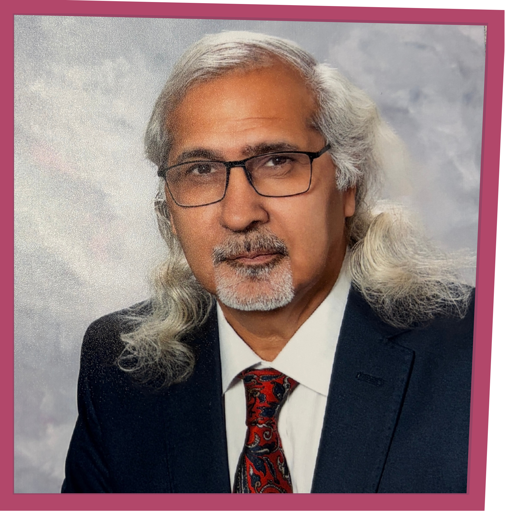 Studio portrait of an older man with long gray hair, glasses, and a gray goatee, wearing a dark suit with a white shirt and red patterned tie.