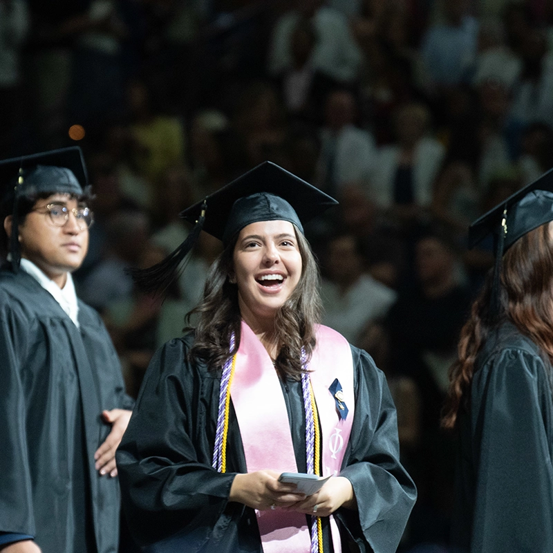 Student in a graduation cap and gown smiles while standing in line at commencement, wearing honor cords and a pink stole, with other graduates nearby.