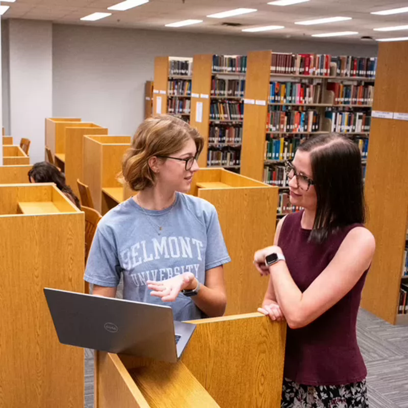 A student standing in the library at a study cubicle with a laptop consulting with the librarian.