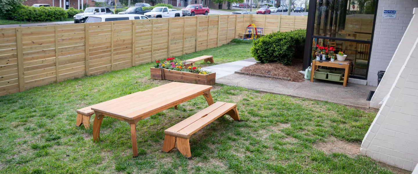 Small outdoor play area with a wooden table, garden boxes and fenced grass yard outside a classroom.