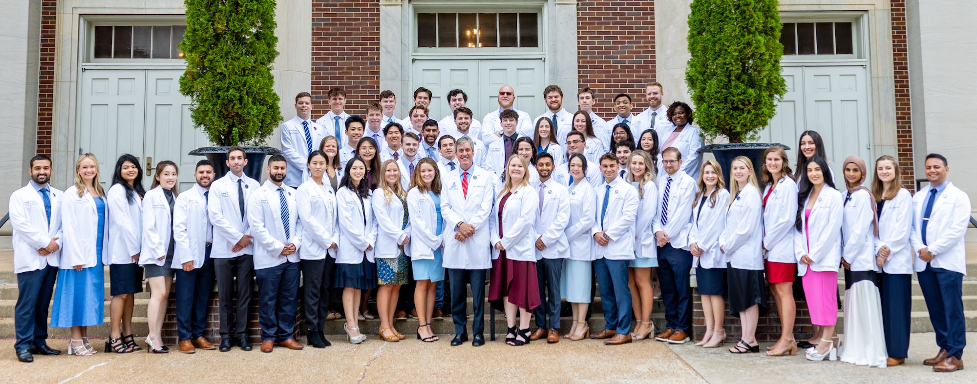 The Thomas F. Frist, Jr. College of Medicine at Belmont University Class of 2029 poses for a group photo on the steps of a campus building. The students, wearing white lab coats, stand together smiling and looking towards the camera. 