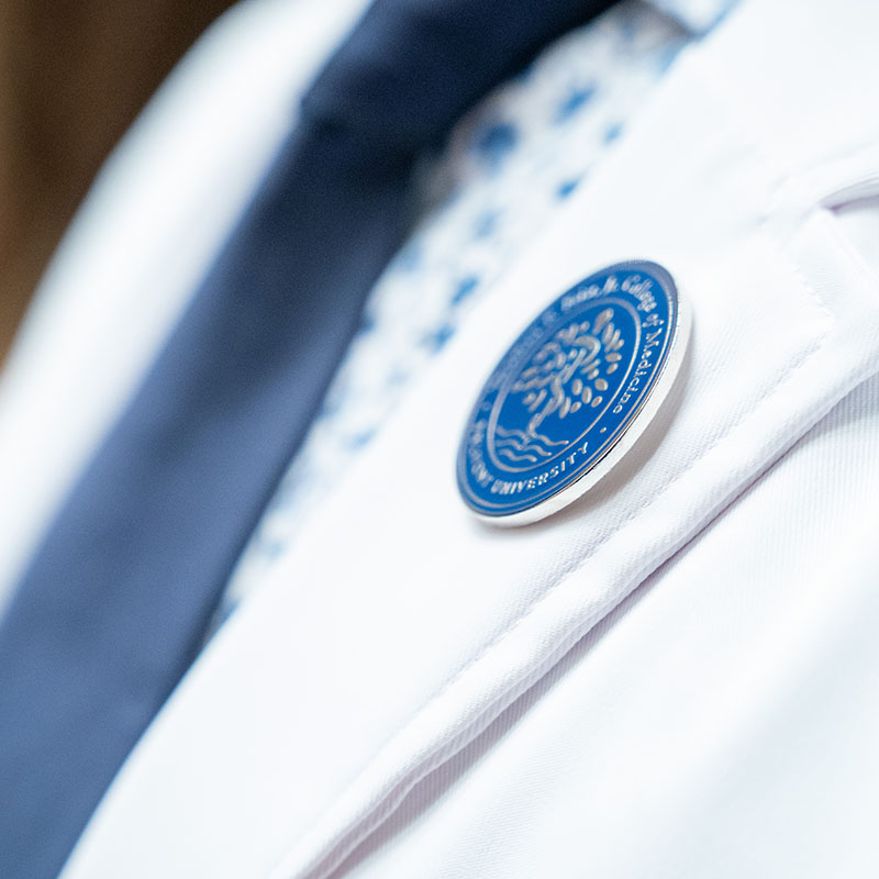 Close-up of a Belmont University medical student's white coat, focusing on a circular blue and white seal of the Thomas F. Frist, Jr. College of Medicine pinned to the coat.