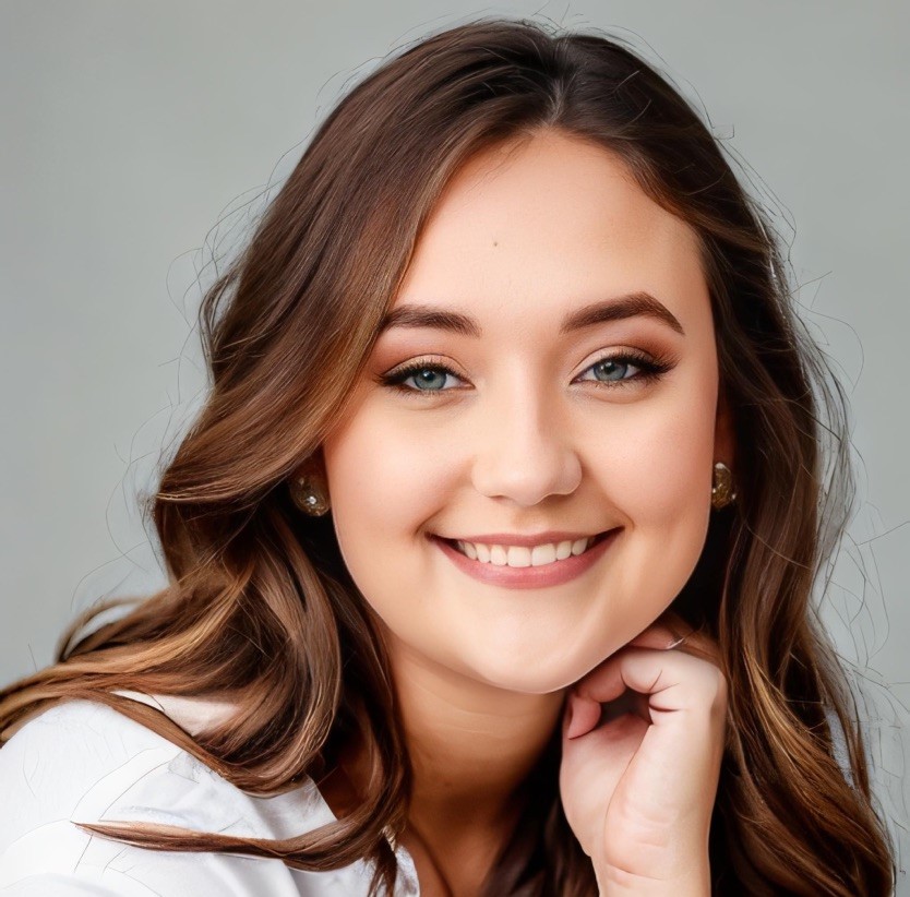 Professional headshot of Amelia Harmond, featuring her with long wavy brown hair, blue eyes, and a warm smile. She is wearing a white blouse and pearl stud earrings, posed against a neutral gray background.