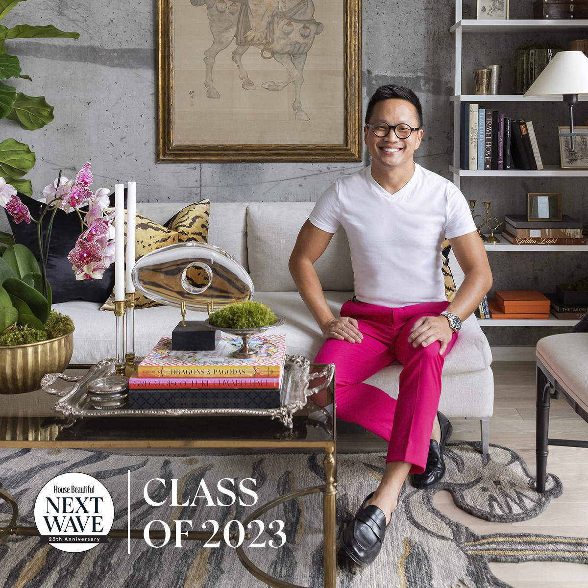Interior designer Evan Millard, wearing a white t-shirt and bright pink pants, sits smiling on a modern light-colored sofa in a stylish, art-filled living room. The space features a decorative coffee table with books, a sculpture, and orchids. 'House Beautiful Next Wave Class of 2023' text appears in the lower left corner.