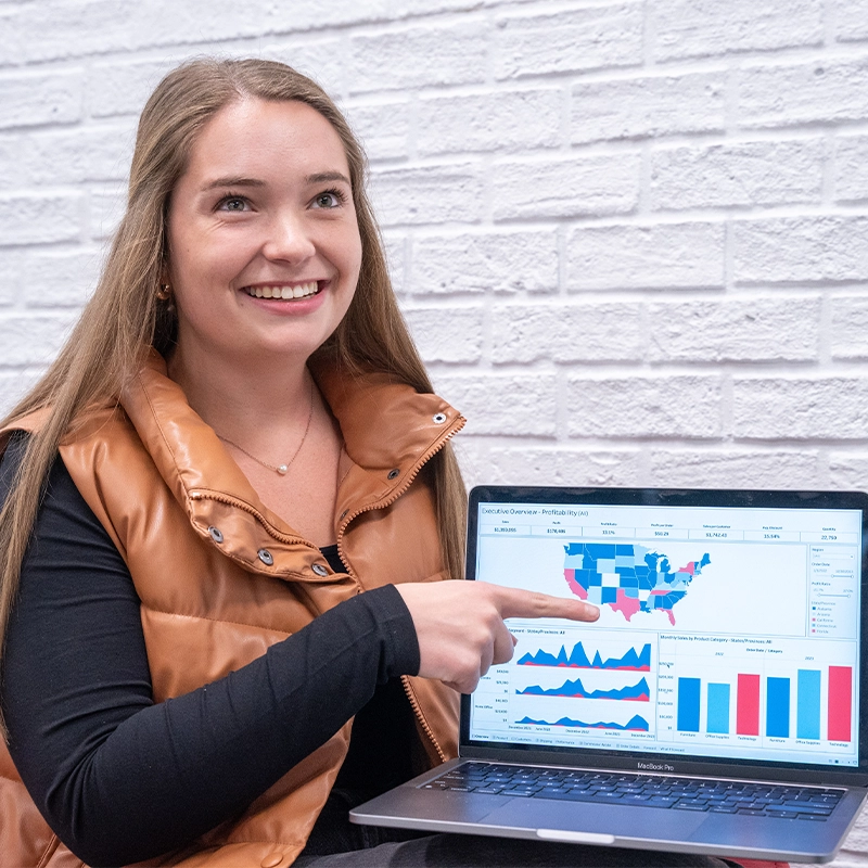 A lady seated indoors, pointing at a laptop screen that has data graphs on it.