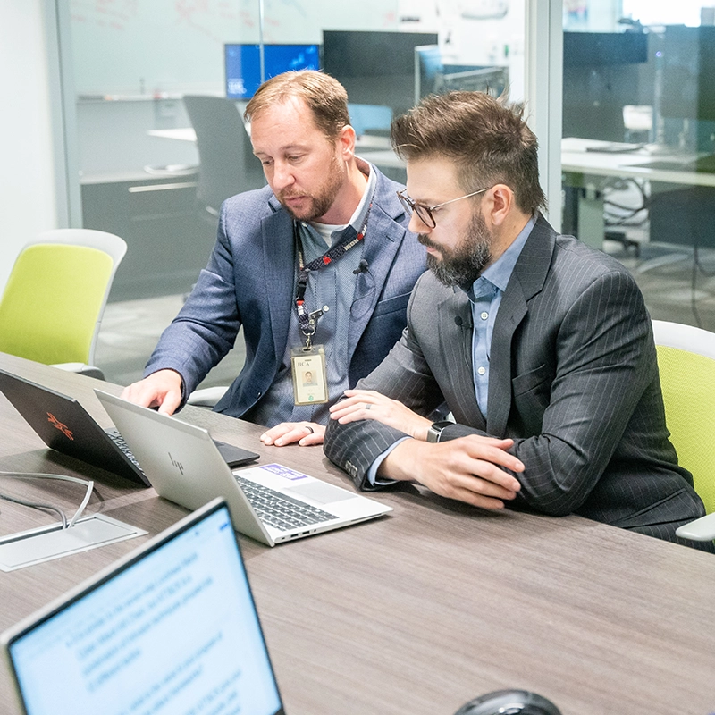 Two men in business suits at a table looking at the same computer.