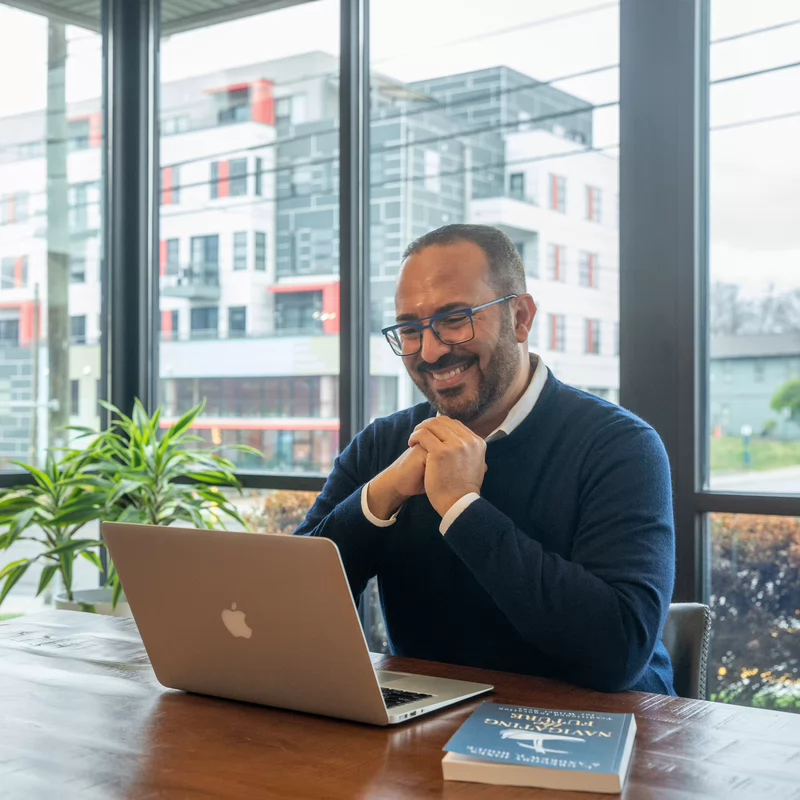 Smiling man with glasses works on a laptop with a book beside it, at a wooden table.