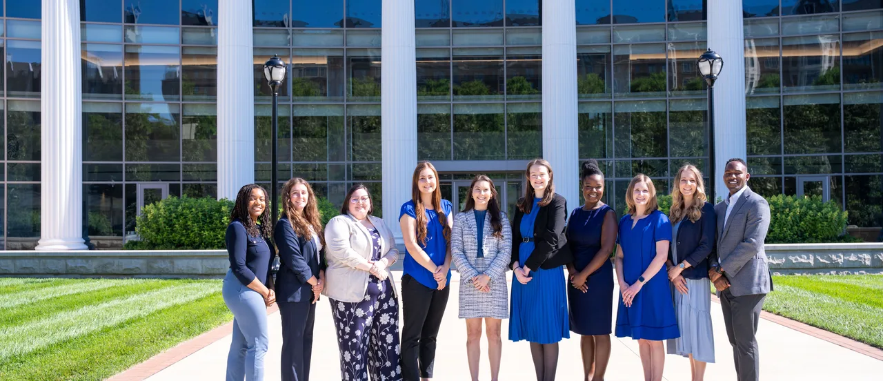 Group photo of 10 diverse business team members, including men and women wearing business attire, posing in front of a contemporary glass-fronted office building with white architectural columns and manicured landscaping
