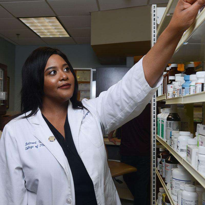 A pharmacy student reaching up to pull a drug container off a shelf