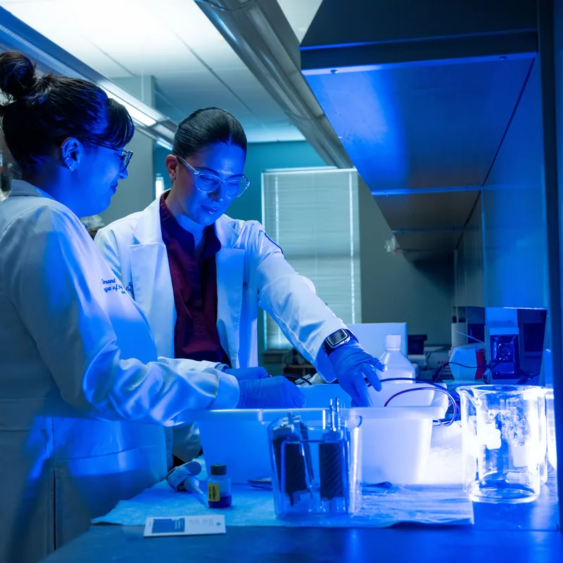 Two pharmacy students wearing safety glasses, white lab coats, and blue nitrile gloves work together at a laboratory bench with glassware and equipment under blue lighting in a modern pharmaceutical laboratory.
