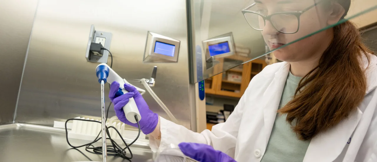 Pharmacy student at Belmont University wearing glasses, a white lab coat, and purple nitrile gloves handles a pipette in a laboratory setting with stainless steel surfaces and computer monitors in the background.