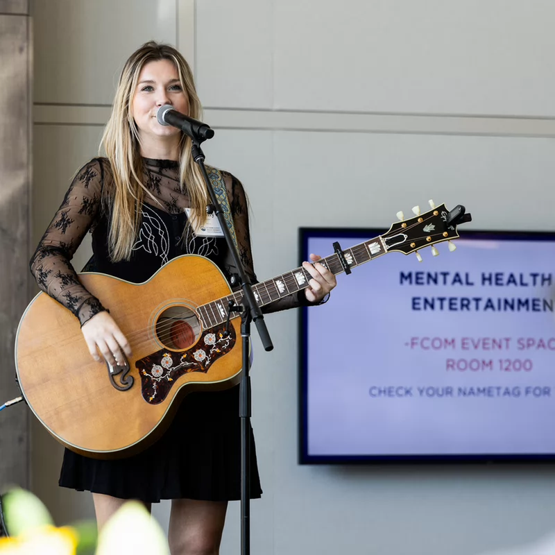 A woman performs on stage with an acoustic guitar, singing into a microphone. She wears a black dress with lace sleeves. A screen behind her reads "Mental Health in Entertainment." 