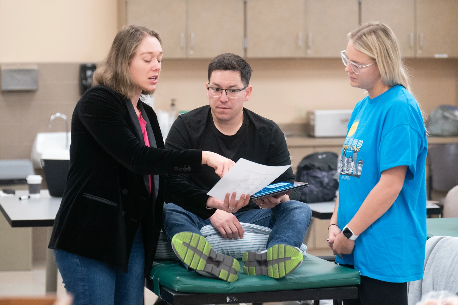 A woman in a black blazer explains a document to a seated man and a standing woman in a blue shirt. They look focused, in a classroom setting.
