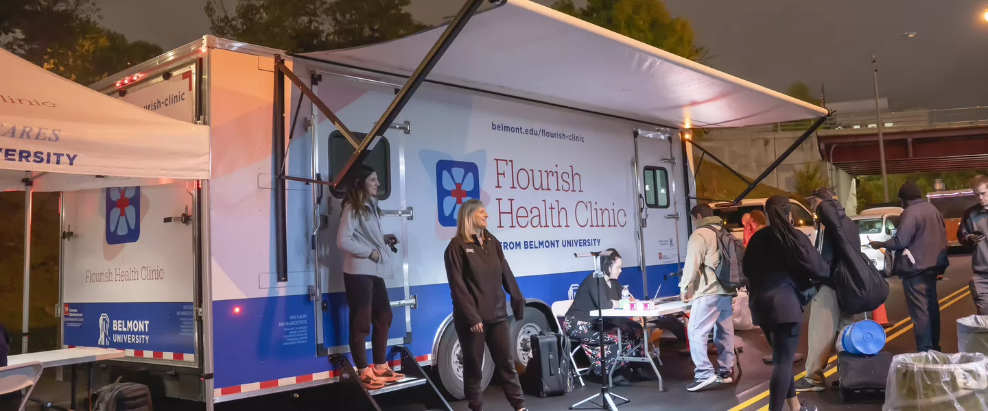 People gather under a tent beside a "Flourish Health Clinic" mobile unit on a street at night. The scene is busy and illuminated by streetlights.
