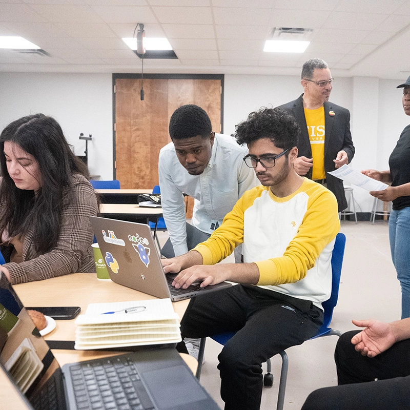 Students collaborate in a classroom setting, working on laptops with focused expressions, while a faculty member in a blazer and yellow shirt engages with another student in the background.
