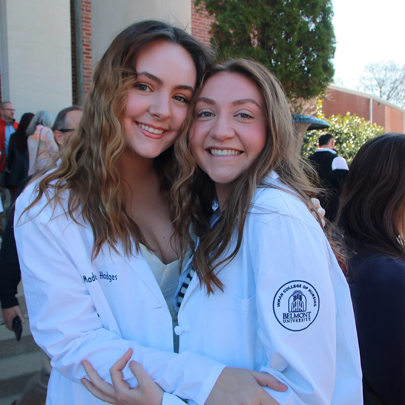Two smiling Belmont nursing students wearing white coats embrace outdoors after a ceremony, with sunlight and other attendees in the background.