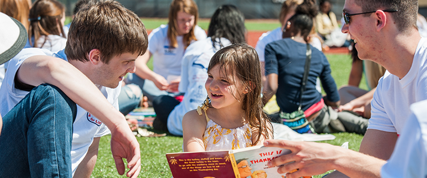 a child sits in the grass and reads with two volunteers