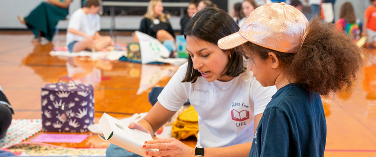 Belmont student is sitting on the floor of a gym reading a book to a young girl