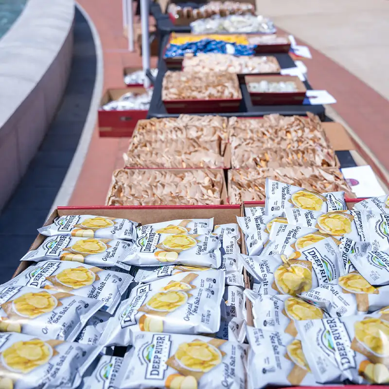 A table is set up outdoors displaying a variety of snacks, including bags of potato chips and other packaged treats. The setup is part of a free lunch event at Belmont University, with a bright and inviting atmosphere.