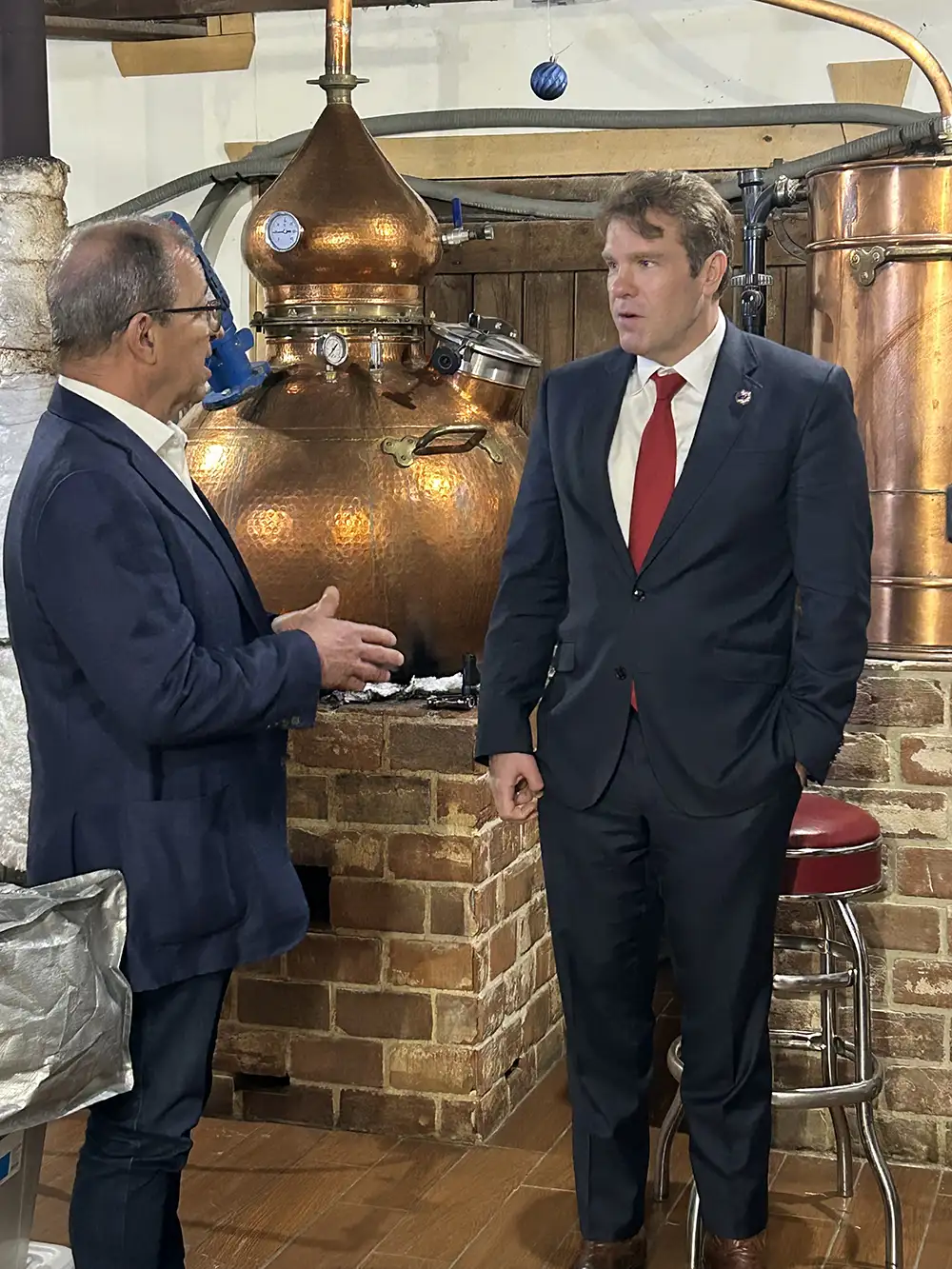 Two men in suits talk beside a large copper distillation still inside a distillery.