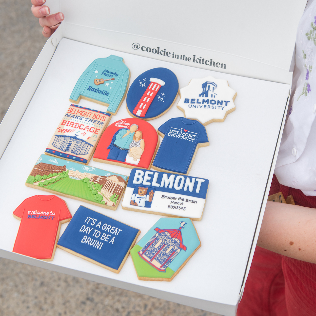 A box of decorative cookies from "@cookie in the kitchen" featuring Belmont University-themed designs. The box contains various cookies shaped like sweaters, t-shirts, and buildings with Belmont University logos, slogans such as "It's a great day to be a Bruin!" and Nashville-related imagery.
