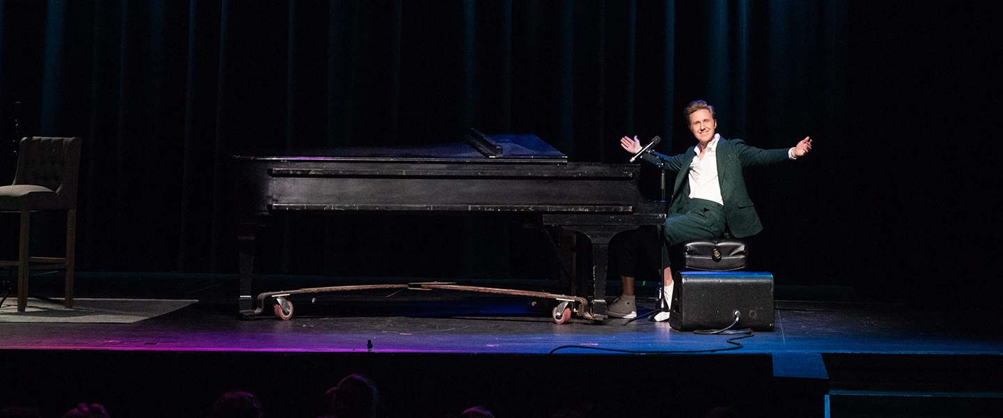 Cody Fry sits at a grand piano on stage, dressed in a green suit with a white shirt. He spreads his arms wide, smiling towards the audience, as if acknowledging applause or engaging with the crowd.