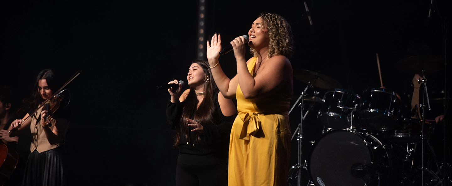 A group of female musicians; one in the background playing violin while two other women are in the foreground singing into handheld microphones