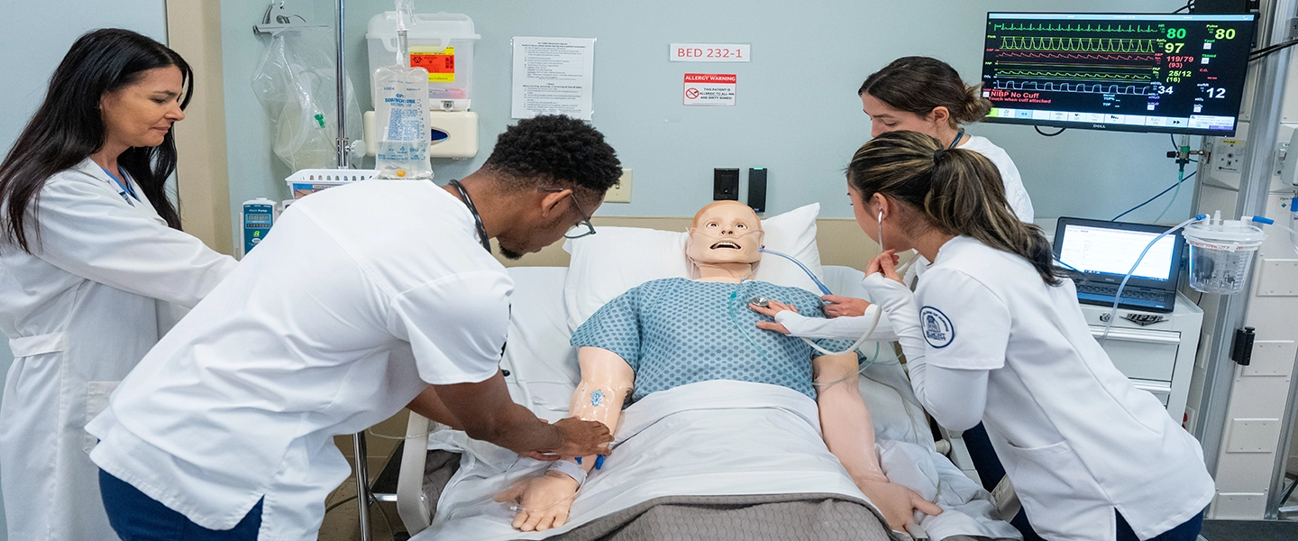 A group of nursing students surrounding a hospital bed and practicing on a practice dummy lying in the bed