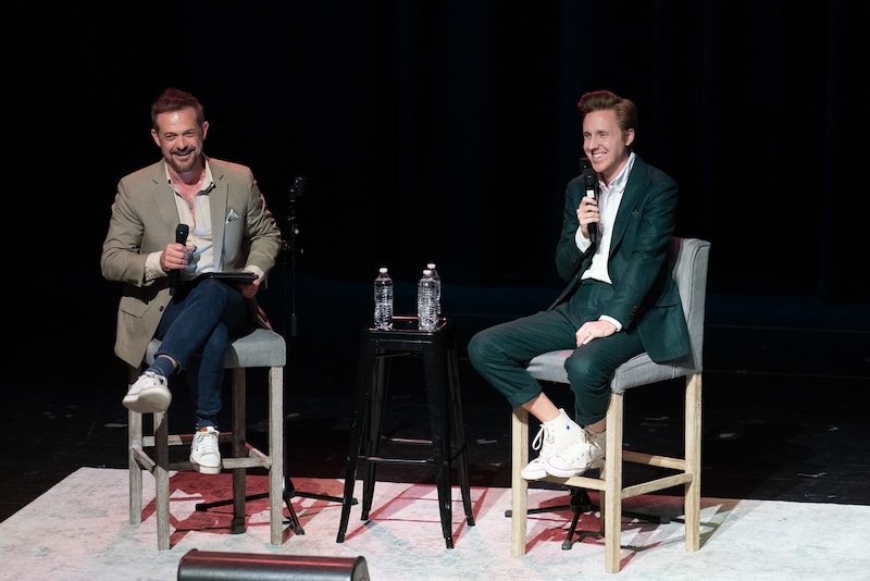 Cody Fry, dressed in a green suit and white sneakers, sits on a tall chair on stage, holding a microphone during a live conversation. Next to him, a host in casual attire with a blazer, jeans, and sneakers also holds a microphone and smiles, engaging with Fry.