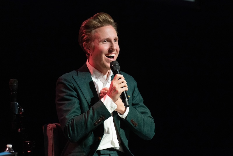 Cody Fry, wearing a green suit and white shirt, smiles warmly while holding a microphone during a live performance. His expression is engaging, as if he is sharing a personal story or interacting with the audience.