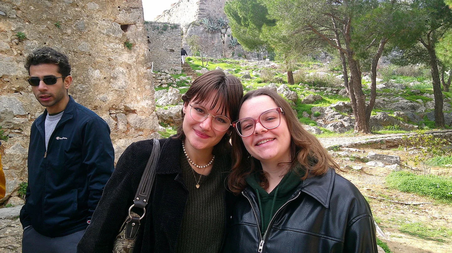 Three students at an ancient ruin site in Athens, Greece. Two students in the foreground smile for the camera while another stands nearby. Ancient stone walls and Mediterranean vegetation visible in the background.