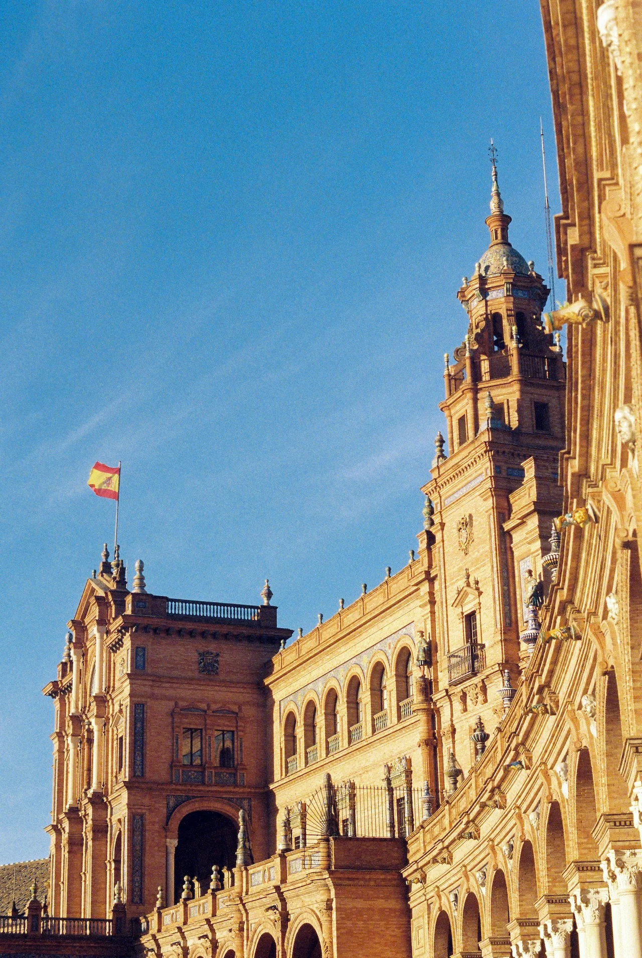 Narrow historic street in Seville, Spain, with a Gothic church tower visible at the end of the perspective. Traditional Spanish architecture lines both sides of the quiet street where cars and motorcycles are parked. The muted, vintage-toned photograph captures the charm of Seville's old town under an overcast sky.