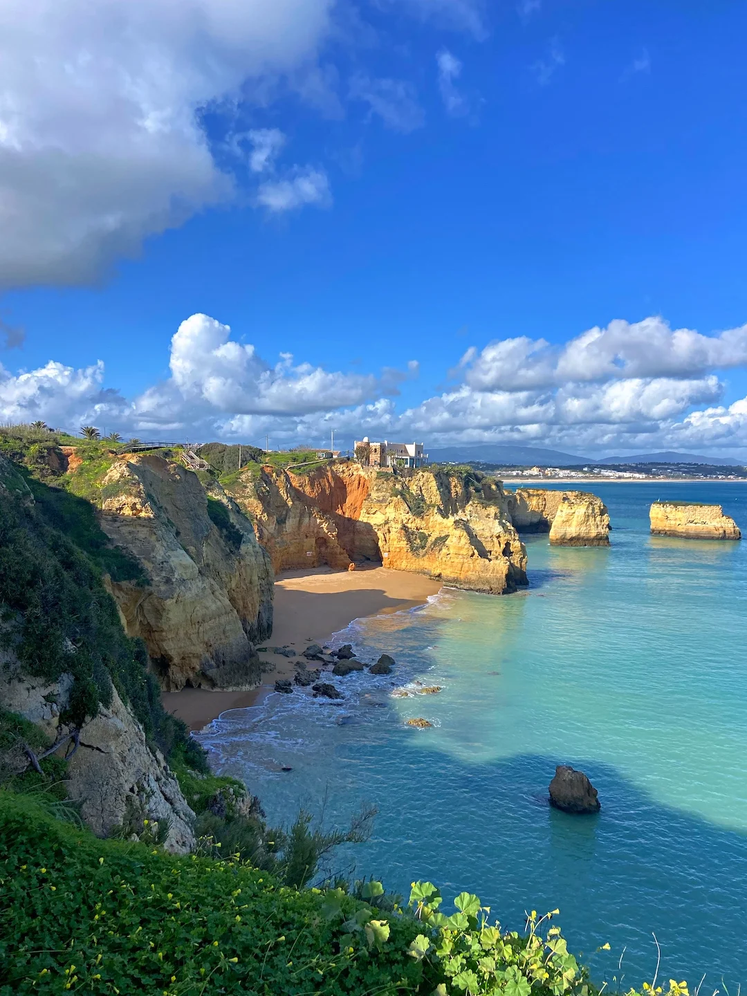Breathtaking coastal landscape at Praia do Camilo in the Algarve region of Portugal, featuring golden limestone cliffs surrounding a secluded beach cove. Crystal-clear turquoise waters lap against the sandy shore, with distinctive rock formations visible in the ocean. Lush vegetation clings to the cliff edges, and buildings perch atop the cliff under a vibrant blue sky with scattered white clouds.