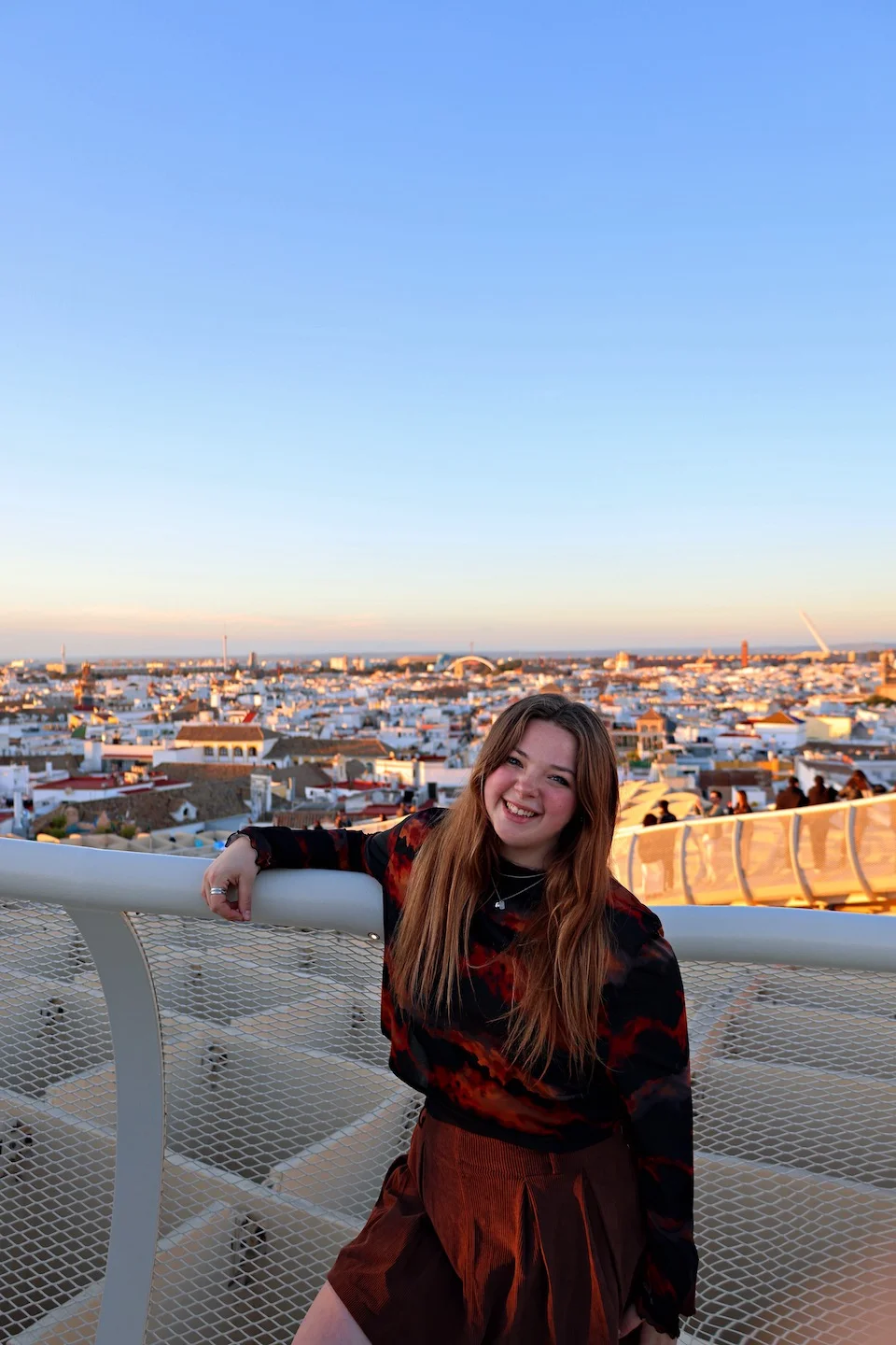 A student smiles at the camera from a viewing platform atop the Metropol Parasol (Las Setas) in Seville, Spain, during sunset. Behind her stretches a panoramic view of Seville's historic white buildings and terracotta rooftops bathed in golden evening light. The modern mesh railing of the observation deck contrasts with the centuries-old cityscape that extends to the horizon under a clear blue sky.