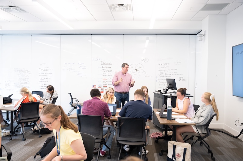 A man in a pink button-up shirt stands and speaks to a classroom of students seated at tables with laptops and notebooks. Behind him, whiteboard walls are covered in handwritten notes and a Venn diagram, and a wall-mounted screen is visible at right.