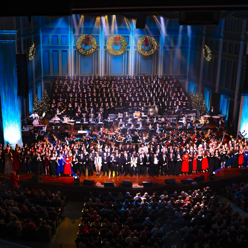 A large ensemble of performers on stage at Christmas at Belmont, featuring a choir and orchestra in a festive venue decorated with Christmas wreaths and lights, filled with an audience.