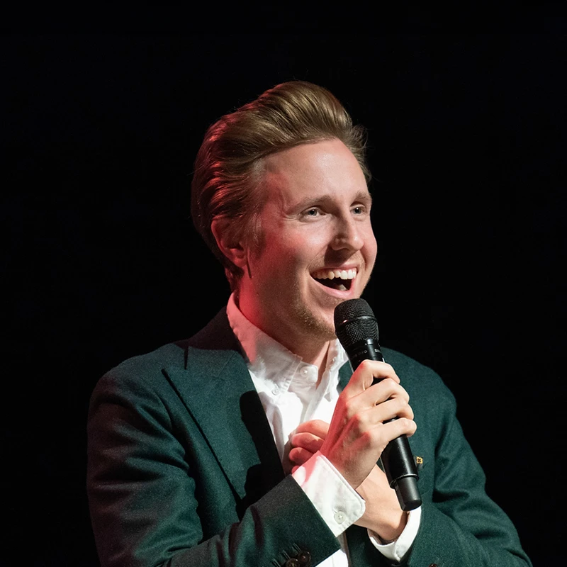 Cody Fry, wearing a green suit and white shirt, smiles warmly while holding a microphone during a live performance. His expression is engaging, as if he is sharing a personal story or interacting with the audience.Alt Text