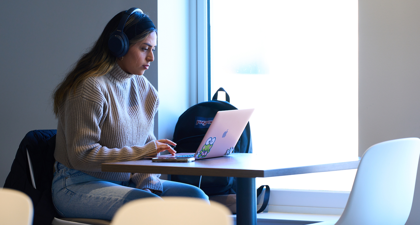 Student studies at a table in a lobby with her laptop and earphones on