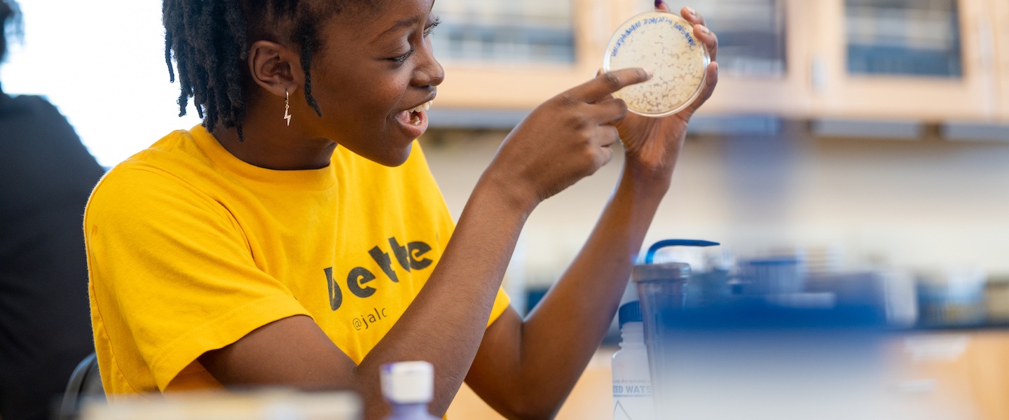 A high school student in a yellow shirt examines a petri dish.