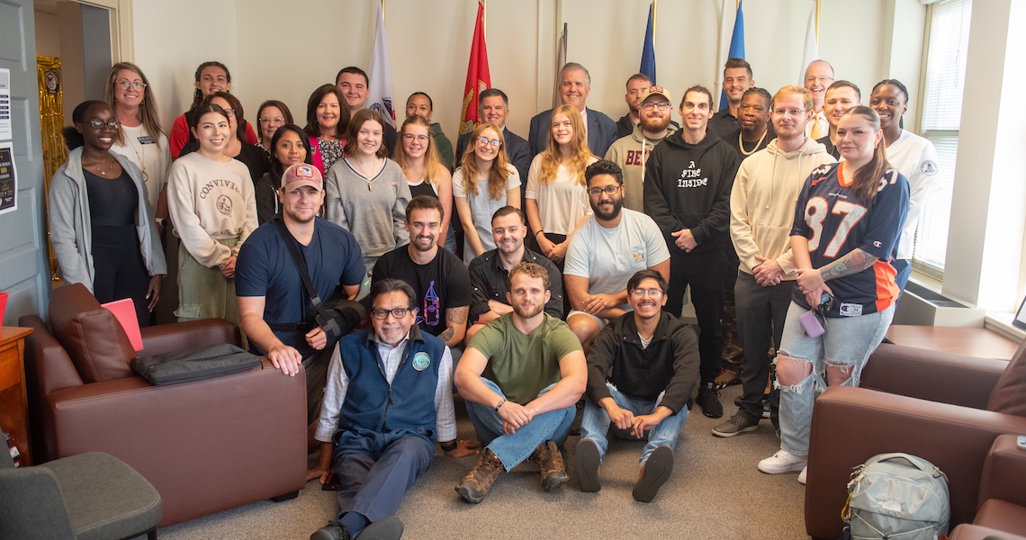 A large group photo of approximately 30 people of diverse ages, genders, and ethnicities in a room. They are arranged in rows, with some sitting on couches in the foreground and others standing behind. The room in the Bruin Vet Center meeting space with flags displayed on the wall in the background.