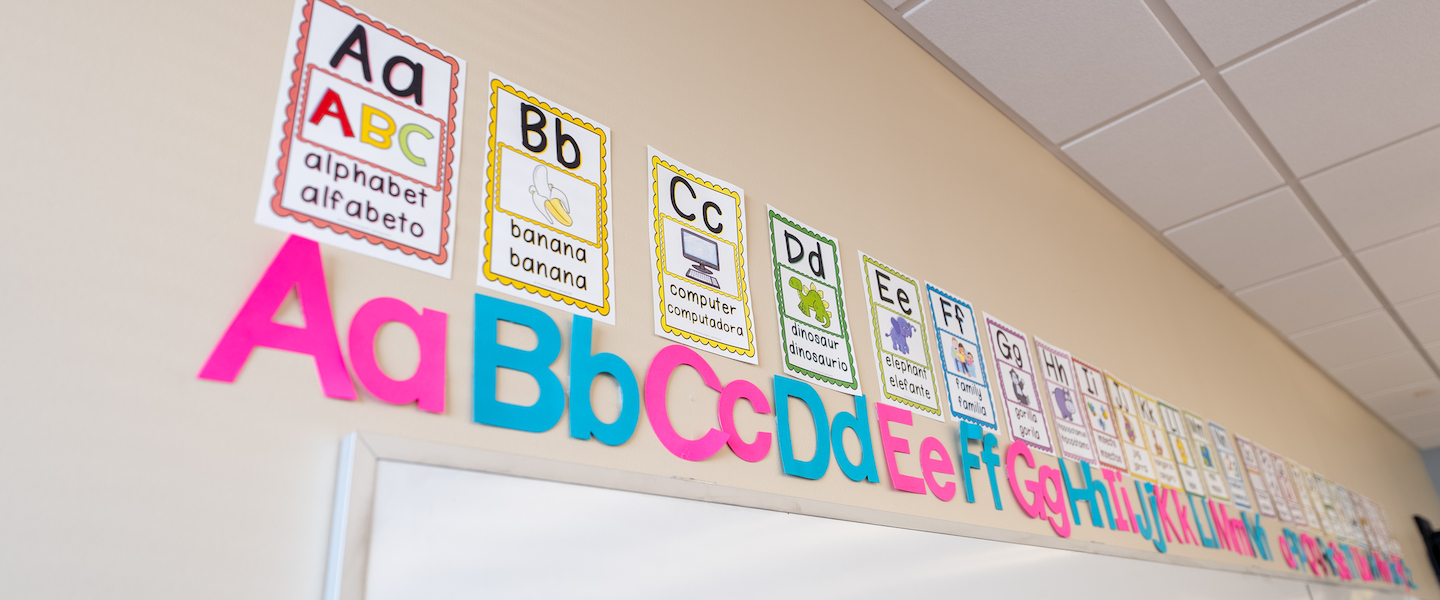 Classroom wall with colorful alphabet display. Large foam letters A-Z below bilingual English/Spanish alphabet flashcards showing letters and corresponding words with images.